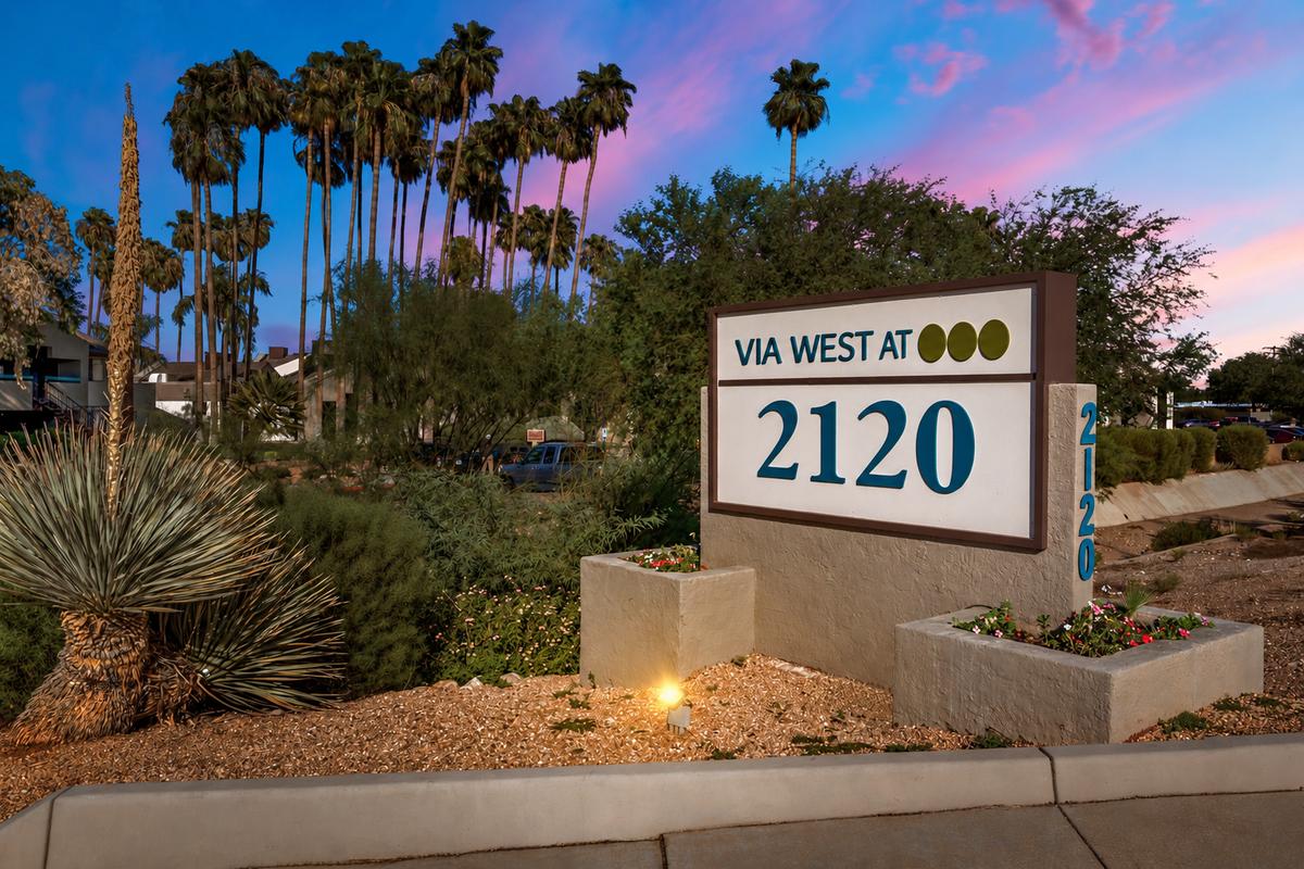 Monument sign at the entrance at dusk, lettered VIA WEST AT 2120 with three olive dots, Tucson palms and a pink sky behind